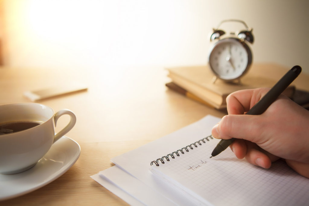 A person writing on a notebook with a clock and hot beverage nearby on a table. 