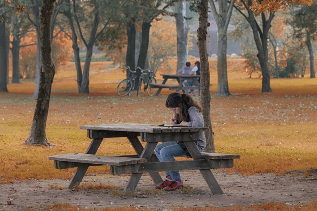 A young woman writing while sitting on a bench at a park.