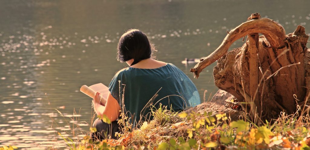 A young woman reading a book near a pond. 