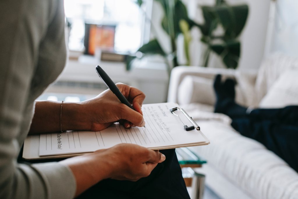 A woman writing notes from a clipboard as another person lies on a couch nearby. 