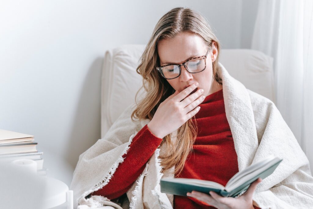 A young woman yawning while reading a book. One of the 6 pitfalls to avoid when writing a young adult novel is not writing flat characters.