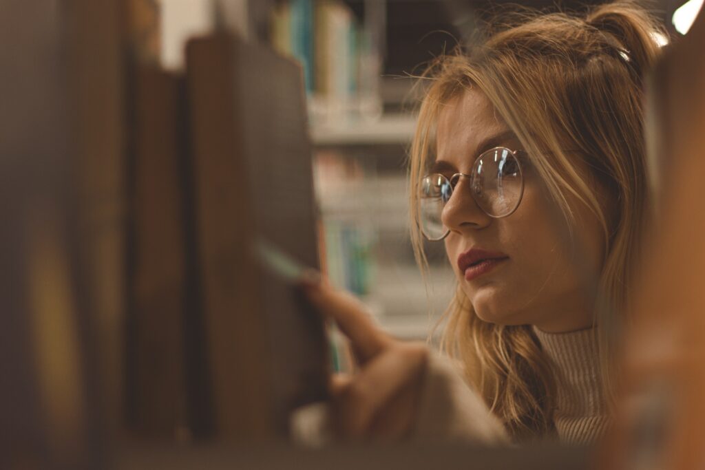 A young woman looking at books at a bookshelf. One of the 6 pitfalls to avoid when writing a young adult novel is not reading books.