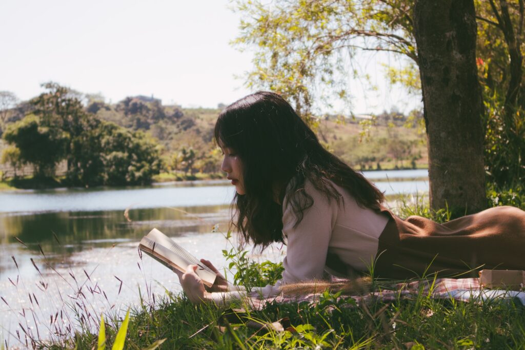 A young girl reading a book on the grass near a pond. 