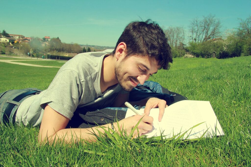 A man busily writing his hook in his notebook while laying on the grass. 