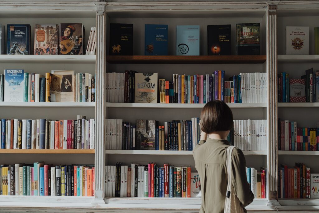 A woman standing in front of a book shelf. Reading books is one way to improve your world-building for YA fiction.