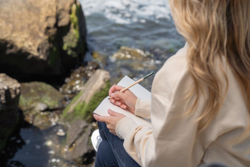 A woman sitting by the water and writing in a journal.