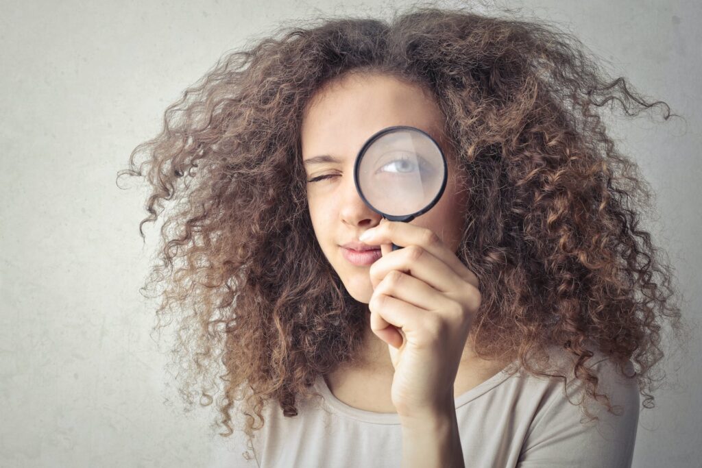 A woman holding up a magnifying glass over her eye. Foreshadowing is a part of mastering plot twists in YA novels. 